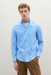 Camicia celeste con bottoni, maniche lunghe, taschino sul petto e orlo leggermente curvo, abbinata a pantaloni marroni. Texture liscia.