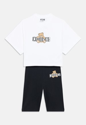 Bela bombažna majica z grafiko medveda in napisom "MOSCHINO", v kombinaciji s črnimi bombažnimi kratkimi hlačami z podobno medvedjo zasnovo na strani.
