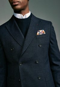 Mörkblå dubbelknäppt blazer i texturerad tyg, med svarta knappar. Lagt över en mörkstickad tröja och vit skjorta med krage. Ficknäsduk med geometriskt mönster.