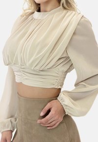 Beżowy krótki top z marszczonym materiałem, długimi transparentnymi rękawami i wysokim dekoltem. Tekstura jest gładka z lekkim połyskiem, ukazując pionowe plisowania.