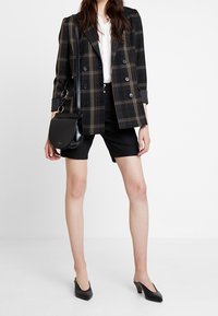 Dwurzędowa marynarka w kratę w kolorze czarnym i beżowym, zestawiona z czarnymi dopasowanymi szortami, czarnymi szpilkami i małą czarną torebką na ramię.