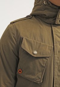 Gros plan sur l'épaule d'une veste verte avec une poche à rabat boutonnée, un œillet marron, et une capuche ajustable avec cordons à bascule.