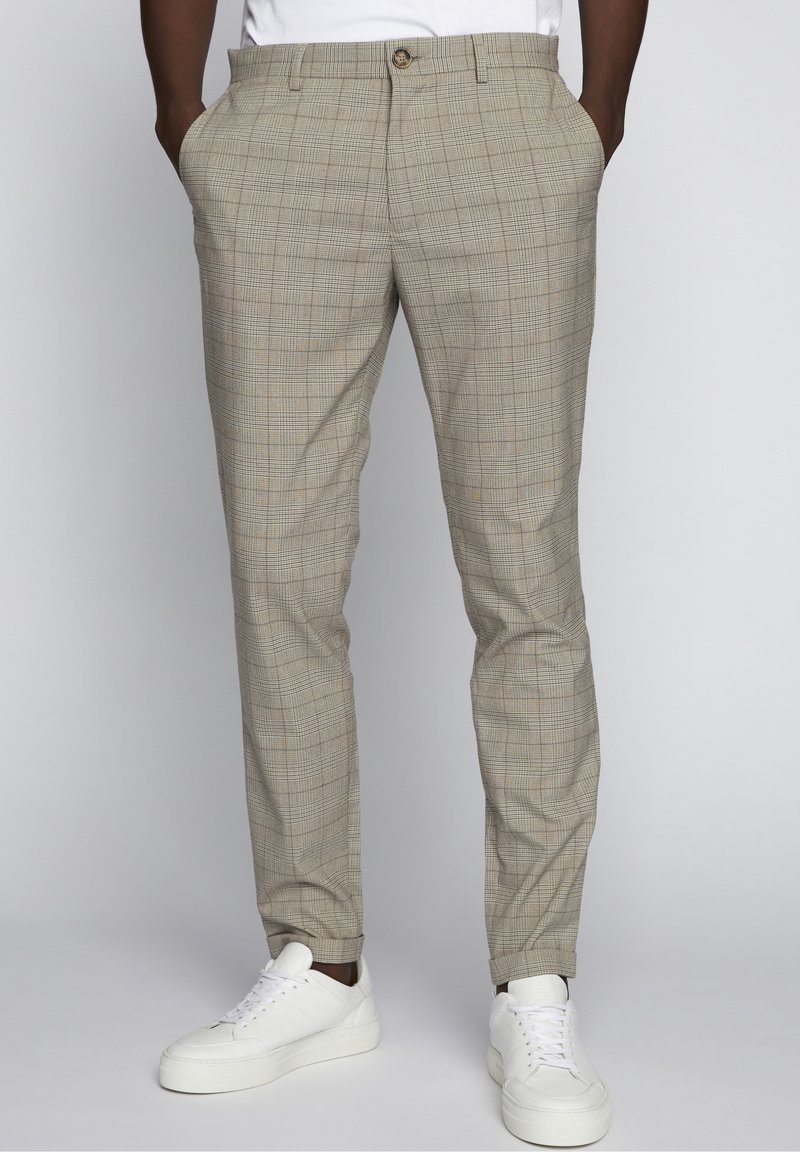 Homme portant un pantalon slim beige à carreaux, les mains dans les poches, et des baskets blanches décontractées sur un fond uni.