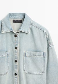Chemise en jean bleu clair avec col, quatre boutons noirs sur le devant et deux poches poitrine, étiquetée Massimo Dutti, fabriquée en Turquie.