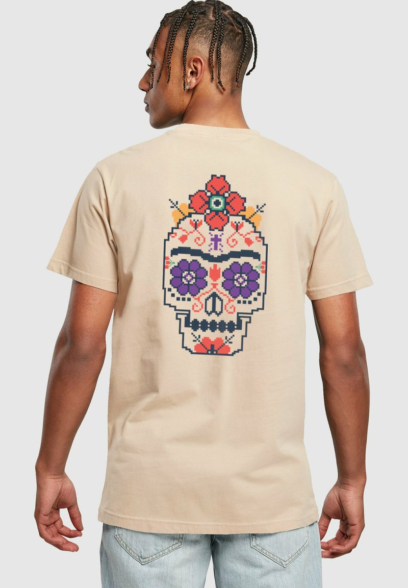 Beige bomull t-skjorte med et fargerikt pixelkunst hodeskalle-design på baksiden, med blomsteraksenter i lilla og røde toner.