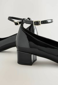 Schwarze Lackleder-Pumps mit quadratischer Spitze, einem mittelhohen Blockabsatz und einem Knöchelriemen mit einer metallischen Schnalle.
