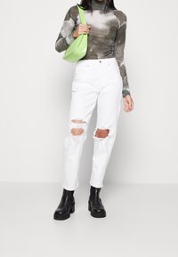 Bílé roztrhané džíny, dlouhý šedý tie-dye top a černé masivní kotníkové boty. Zelená kabelka přehozená přes rameno. Minimalistické pozadí.