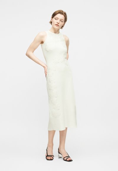 Γυναίκα που φοράει λευκό αμάνικο midi φόρεμα με κεντήματα λουλουδιών και μαύρα τακούνια με λουράκια, όρθια με τα χέρια πίσω από την πλάτη.