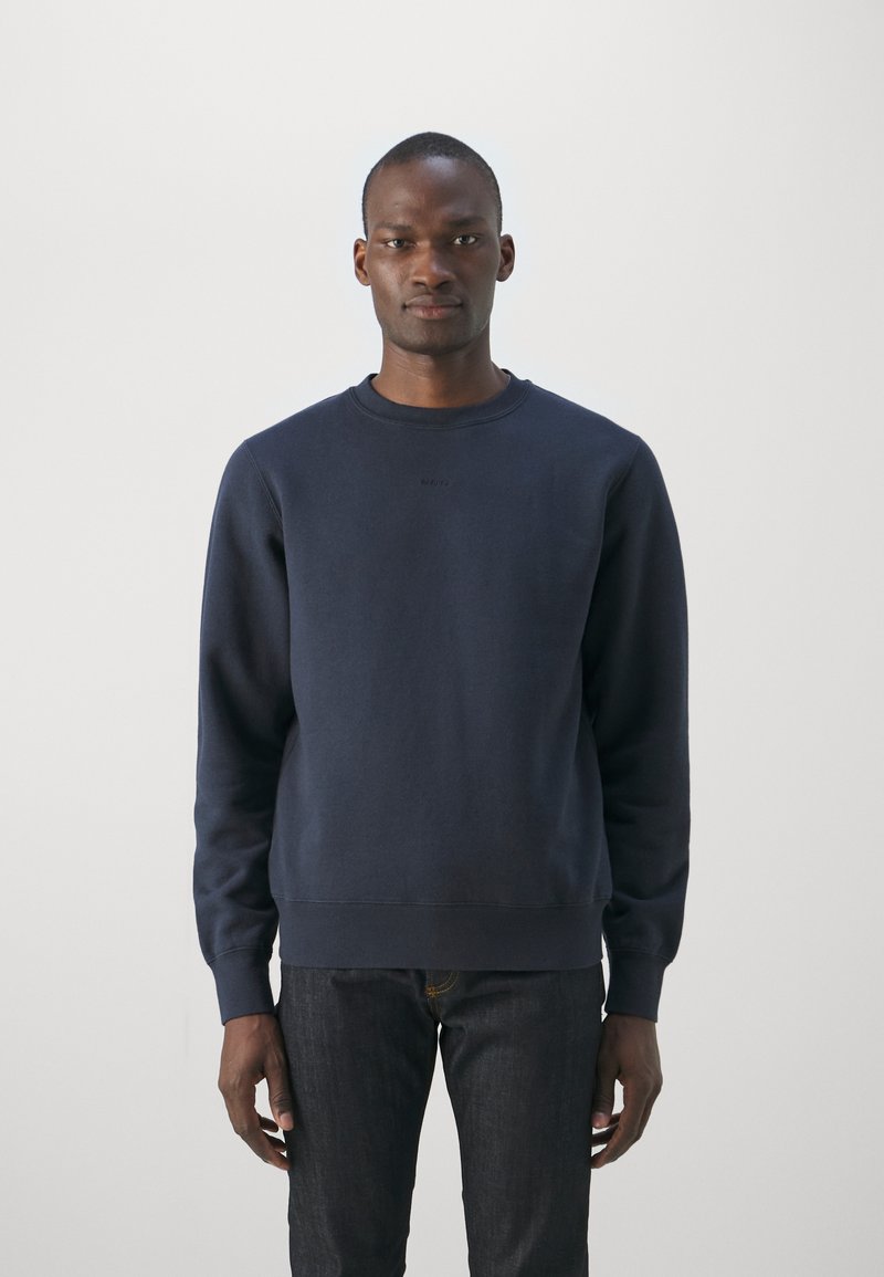 Navyblauer Pullover aus Baumwolle mit Rundhalsausschnitt, langen Ärmeln sowie gerippten Bündchen und Saum. Kombiniert mit dunklen Jeans.