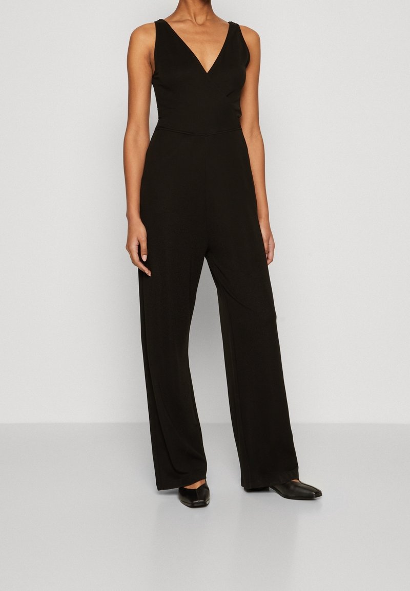 Femme portant une combinaison noire sans manches à jambes larges, avec un col en V et des chaussures plates noires, debout sur un sol gris uni.