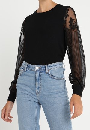 Top en tricot noir avec des manches en dentelle rayée transparente ornées de détails floraux, associé à un jean en denim taille haute bleu clair.