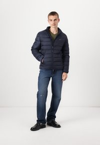Marineblauwe gewatteerde jas met een rits aan de voorkant, voorzien van een staande kraag, horizontale quiltstiksels en twee zijzakken. G gedragen over een groene shirt en spijkerbroeken.