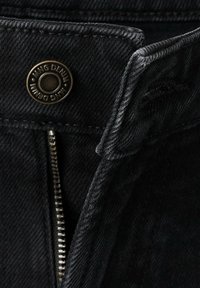 Gros plan d’un jean en denim noir dézippé montrant les dents de la fermeture éclair et un bouton métallique portant l’inscription "MNG DENIM."