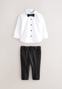 Chemise blanche à manches longues avec boutons noirs et nœud papillon, associée à un pantalon noir sur mesure sur un fond neutre.
