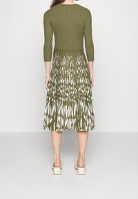 Robe tricotée vert olive avec un corsage côtelé et une jupe évasée mi-longue ornée d'un motif abstrait blanc. Manches mi-longues, taille cintrée.
