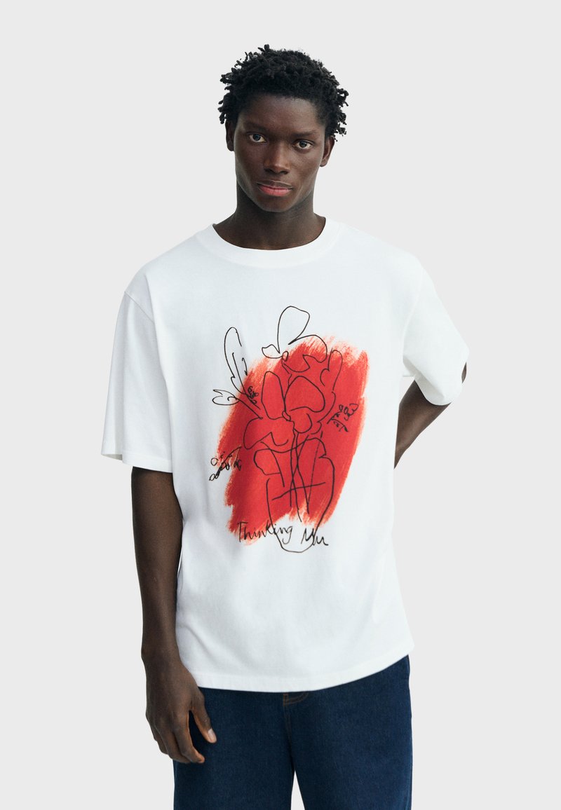 Jeune homme portant un T-shirt blanc oversize avec une fleur abstraite rouge et le texte "Thinking Me", assorti à un jean bleu foncé.