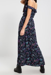 Robe maxi fleurie décolletée noire avec motifs violet et sarcelle, dotée d'une taille cintrée et d'une jupe évasée. Portée avec des bottes noires.