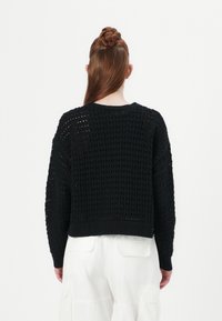 Pull en tricot noir avec un motif de tissage ouvert. Il présente un col rond et des manches à bords côtelés au niveau de l'ourlet et des poignets.