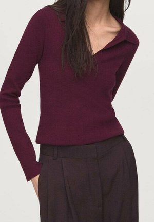 Vrouw draagt een donkerpaarse gebreide top met lange mouwen, een brede kraag en een hooggesneden, donkerbruine geplooide broek.