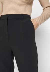 Pantaloni neri su misura in tessuto liscio, con tasche laterali e una vita pulita, abbinati a un top a coste di colore chiaro.