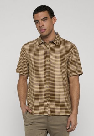 Mężczyzna w brązowej kraciastej koszuli na krótki rękaw z kołnierzykiem i beżowych spodniach, stojący z jedną ręką w kieszeni na szarym tle.