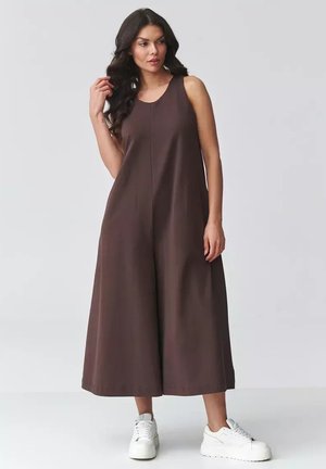 Kobieta w brązowym kombinezonie bez rękawów o szerokich nogawkach, w białych trampkach, stojąca na tle jednolitego jasnoszarego tła.