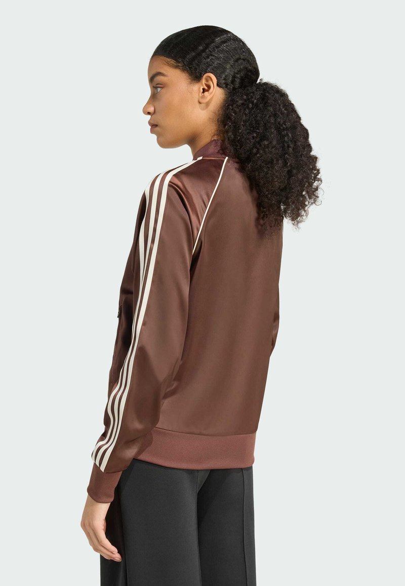 Brown satin bomber jacket with white stripes along the sleeves, fitted cuffs, and a high collar, paired with black pants.