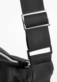 Zwarte stoffen tas met een dikke, gevlochten riem met een zilveren metalen gesp en een zichtbare rits langs de bovenrand.