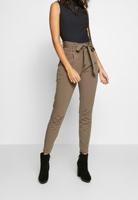 Pantalons marron taille haute avec une taille ceinturée, fabriqués à partir d'un tissu lisse. Associés à un haut sans manches noir à côtes et des bottines noires.