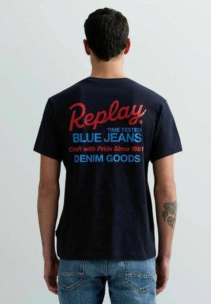 Uomo con capelli corti e scuri e tatuaggio sull'avambraccio sinistro indossa una maglietta nera con scritte rosse e blu e jeans blu, volto di spalle rispetto alla fotocamera.