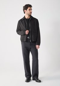 Chaqueta ligera negra con cremallera, cuello y bolsillo delantero. Combinada con pantalones oscuros y zapatos negros, creando un aspecto simple y elegante.