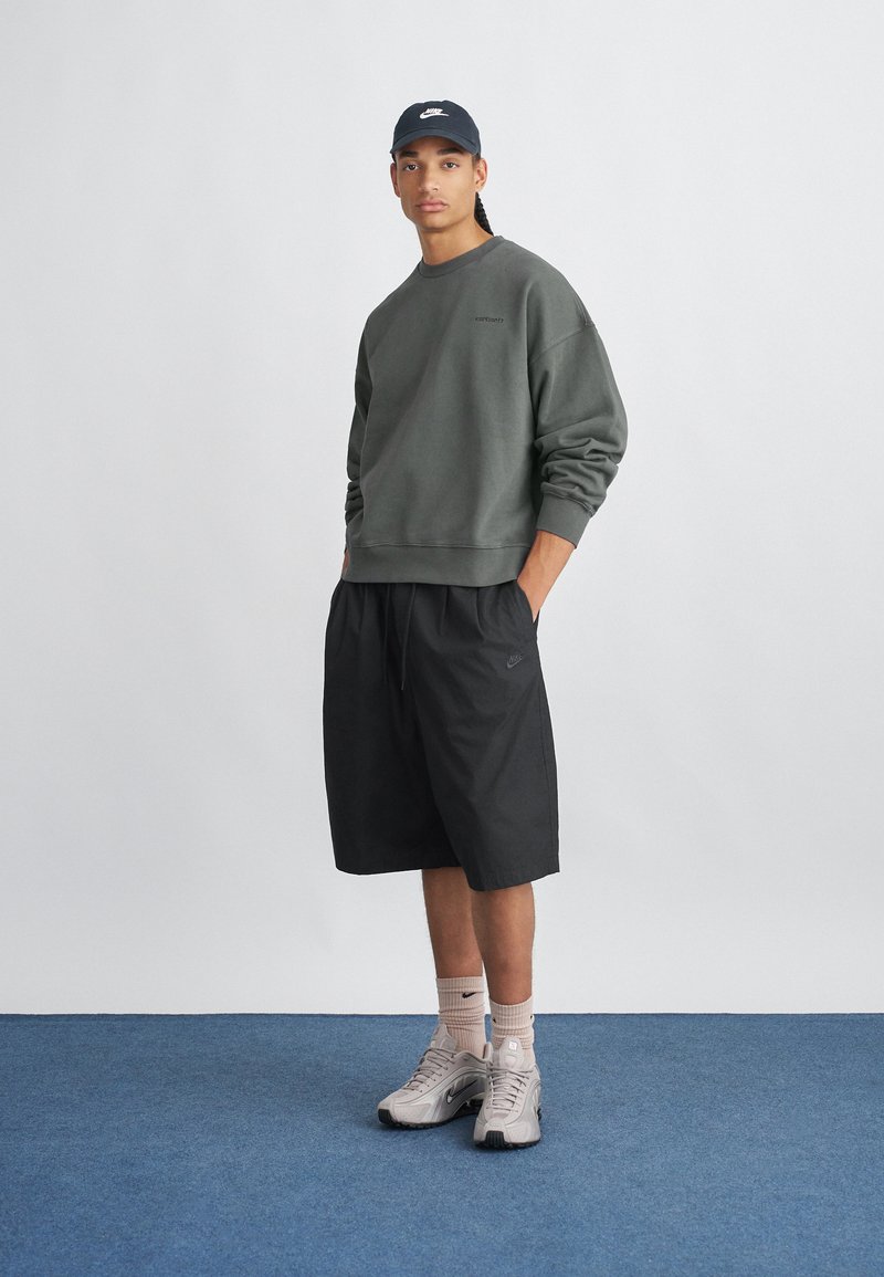 Sudadera verde con puños acanalados, pantalones cortos negros con cordón y zapatillas deportivas gris claro. Gorra azul marino con logo. Fondo liso.
