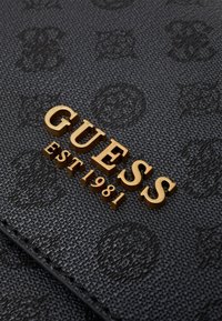 Schwarzes, strukturiertes Material mit eingeprägtem Muster; zeigt das goldene "GUESS"-Logo und "EST 1981" in kontrastierender goldener Schrift.