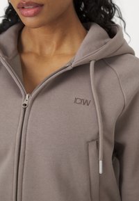 Femme portant un sweat à capuche taupe zippé avec cordons et petit logo brodé sur la poitrine, lèvres et cou visibles.