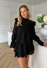 Musta kerroksellinen mekko, jossa on frillikaulus ja hihat, sisältäen monimutkaisia kukkakuosisia yksityiskohtia. Läpinäkyvä kangas laskeutuu pehmeästi, yhdistettynä vaaleaan taustaan.