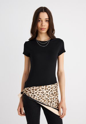 Ung kvinde iført en sort tætsiddende t-shirt, lagdelte sølvhalskæder og et beige leopardprint tørklæde bundet omkring taljen.