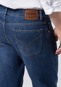 Jeans in denim Brax di colore blu scuro con toppa in pelle marrone chiaro. Caratterizzati da una tasca posteriore con cuciture decorative a forma di X.