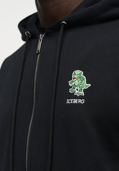 Μαύρο φούτερ με φερμουάρ, με πράσινο γραφικό δεινόσαυρου και κεντημένο το κείμενο "ICEBERG" στο αριστερό μέρος του στήθους.