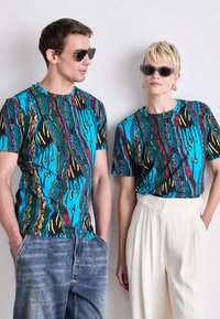 Farverige T-shirts med korte ærmer med abstrakte mønstre i blå, sort, rød og gul, båret af to modeller med forskellige solbriller.