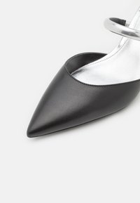 Chaussure en cuir noire à bout pointu avec une semelle intérieure argentée brillante et une bride de cheville argentée métallique sur fond blanc.