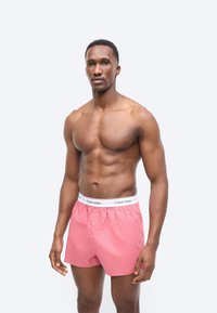 Muskularny mężczyzna z nagim torsen, ubrany w różowe bokserki Calvin Klein, stojący na tle jednolitego jasnoszarego tła.