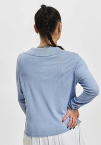 Person med flette i håret, kledd i en lys blå langermet topp og hvit skjørt, med hånden hvilt på hoften, vist fra baksiden.