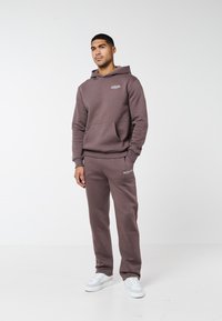 Barna kapucnis pulóvert és hozzáillő szabadidőnadrágot viselő férfi fehér tornacipőben, fehér háttér előtt áll.