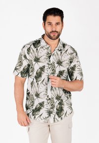 Camisa de botones de manga corta en beige claro con estampado de hojas de palma verdes. Presenta un cuello, corte relajado y textura de tela.