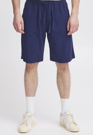 Blue linen shorts with an elastic waistband and drawstring. Features two front pockets and a loose fit, paired with white socks and sneakers.
