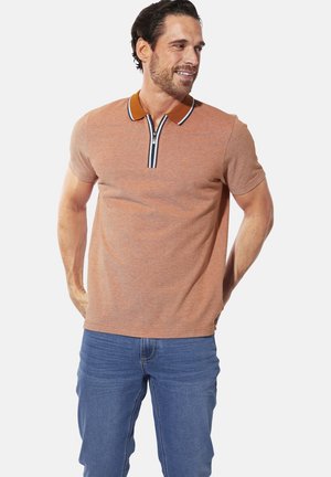 Mann in einem rostfarbenen Poloshirt mit Reißverschlusskragen und blauen Jeans, der mit verschränkten Händen hinter dem Rücken steht, zur Seite schaut und lächelt.