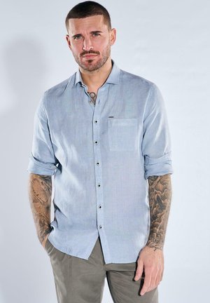 Man with short hair and tattoos wears light blue rolled-up shirt and gray pants, standing with hands in pockets against plain background.
