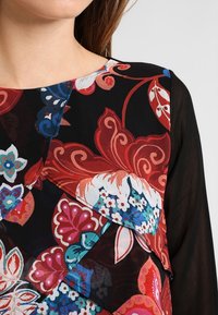 Blouse à motifs floraux avec tissu superposé et vibrant en rouge, rose et bleu sur fond noir. Manches et encolure en voile noir.