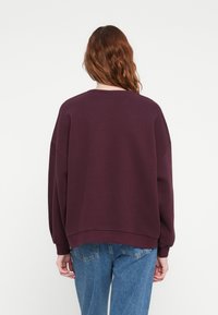 Burgundská oversized mikina s okrúhlym výstrihom, spadnutými ramenami a rebrovanými manžetami, kombinovaná s svetlomodrými džínami. Hladká textúra.