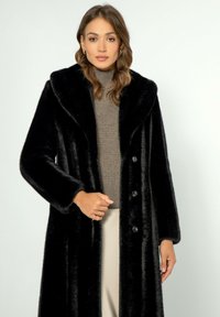 Černý kožich z umělé kožešiny se širokým límcem, třemi knoflíky a dlouhými rukávy, nošený přes hnědý svetr s žebrováním, na světlém pozadí.
